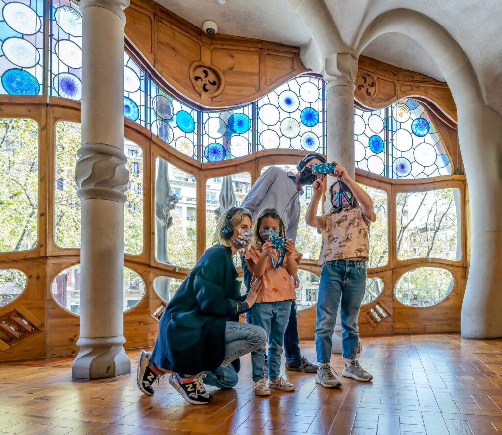 Family wearing masks using audio guides at Casa Batlló, Barcelona. Stained glass windows and unique architecture.
